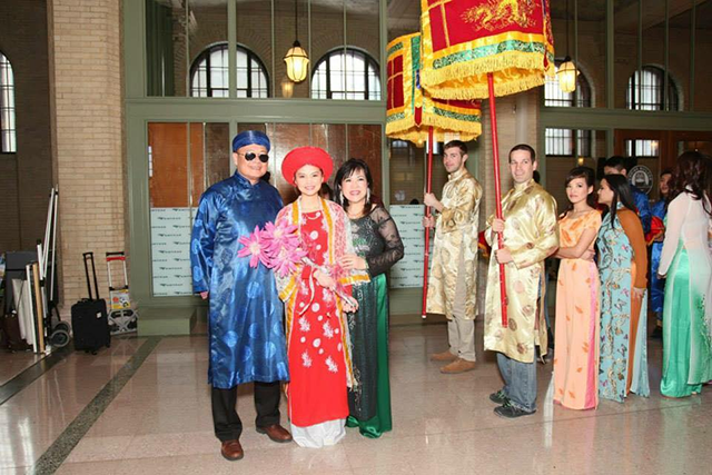 People in traditional Vietnamese dress stand next to a procession of more people led by two leaders holding colorful decorations on poles.